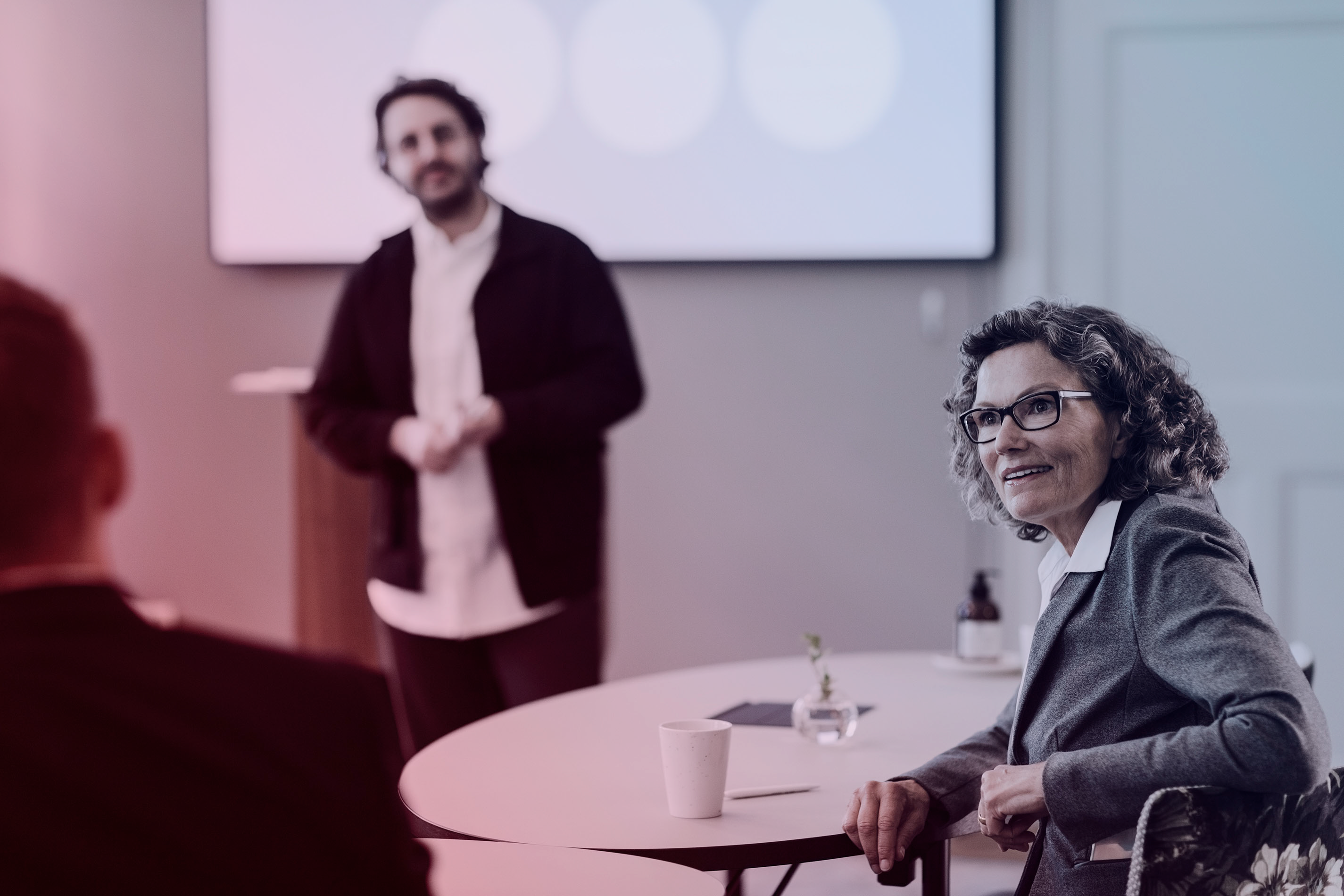 Kvinna i grå kavaj sitter vid ett runt bord och lyssnar engagerat under en presentation, medan en man står framför en skärm i bakgrunden – en scen från ett möte eller seminarium med fokus på arbetsmiljö, ledarskap eller kompetensutveckling.
